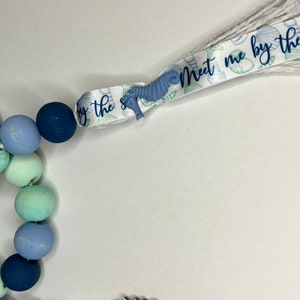 May include: A beaded garland with a coastal theme. The garland features wooden beads in shades of blue and green. A ribbon with the text "Meet me by the sea" and seahorse accents is attached to the garland.