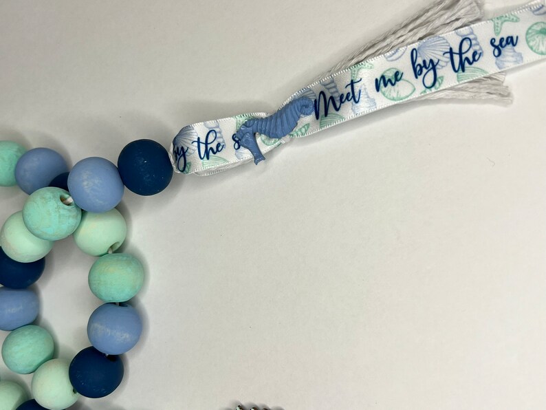 May include: A beaded garland with a coastal theme. The garland features wooden beads in shades of blue and green. A ribbon with the text "Meet me by the sea" and seahorse accents is attached to the garland.