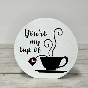 May include: White wooden round sign with black text that reads "You're my cup of" and a black silhouette of a teacup with steam and a teabag with a red heart.