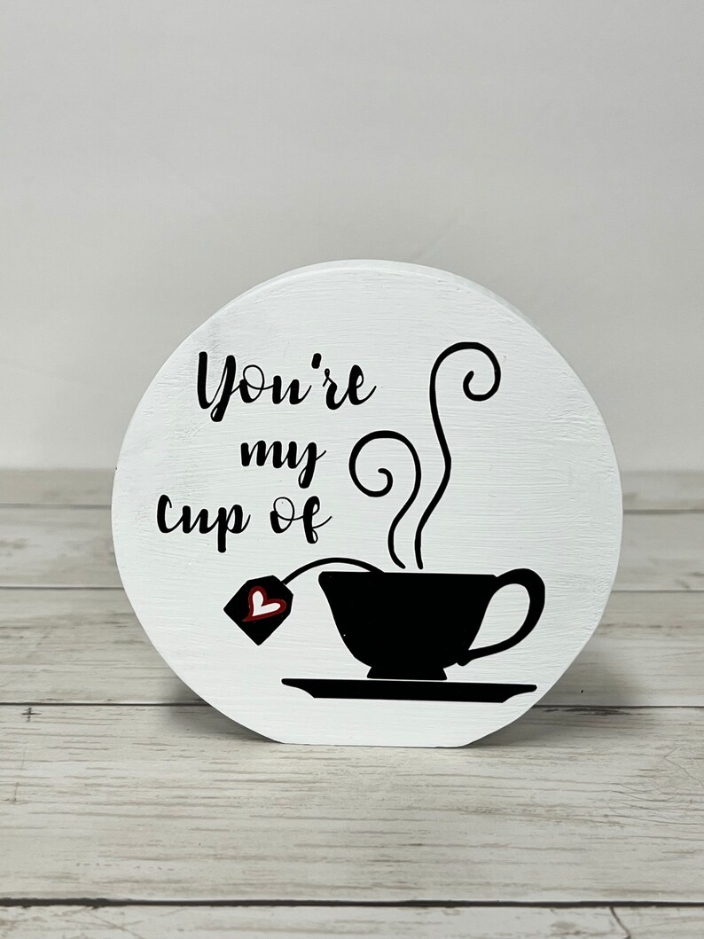 May include: White wooden round sign with black text that reads "You're my cup of" and a black silhouette of a teacup with steam and a teabag with a red heart.