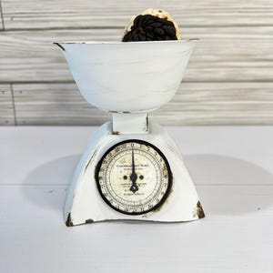 May include: A vintage white scale with a distressed finish, featuring a round dial with black numbers and a black needle. The scale has a bowl containing a dark-colored object. The scale is a decorative kitchen item.