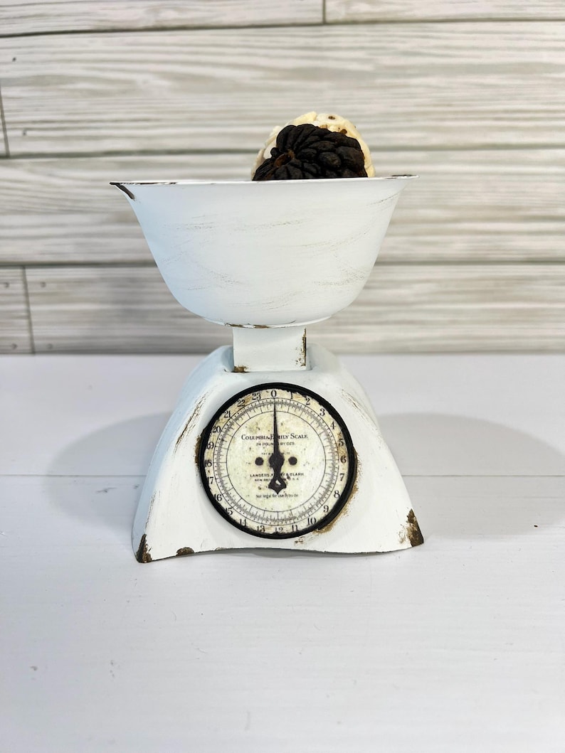 May include: A vintage white scale with a distressed finish, featuring a round dial with black numbers and a black needle. The scale has a bowl containing a dark-colored object. The scale is a decorative kitchen item.