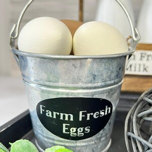 May include: A small, galvanized metal bucket with a silver handle, filled with three eggs. The bucket has a black oval sticker with the words "Farm Fresh Eggs" in white. The eggs are white and brown.