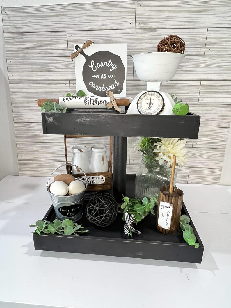 May include: A two-tiered black farmhouse-style decorative tray with various kitchen-themed items. Includes a white scale, a sign that says "Country as cornbread kitchen", a rolling pin, eggs, milk bottles, and greenery.