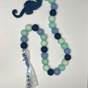 May include: A decorative wooden bead garland in shades of blue and green, featuring a navy blue seahorse cutout. A ribbon with the text "Meet me by the sea" is attached to the garland. The beads are approximately 1 inch in diameter.