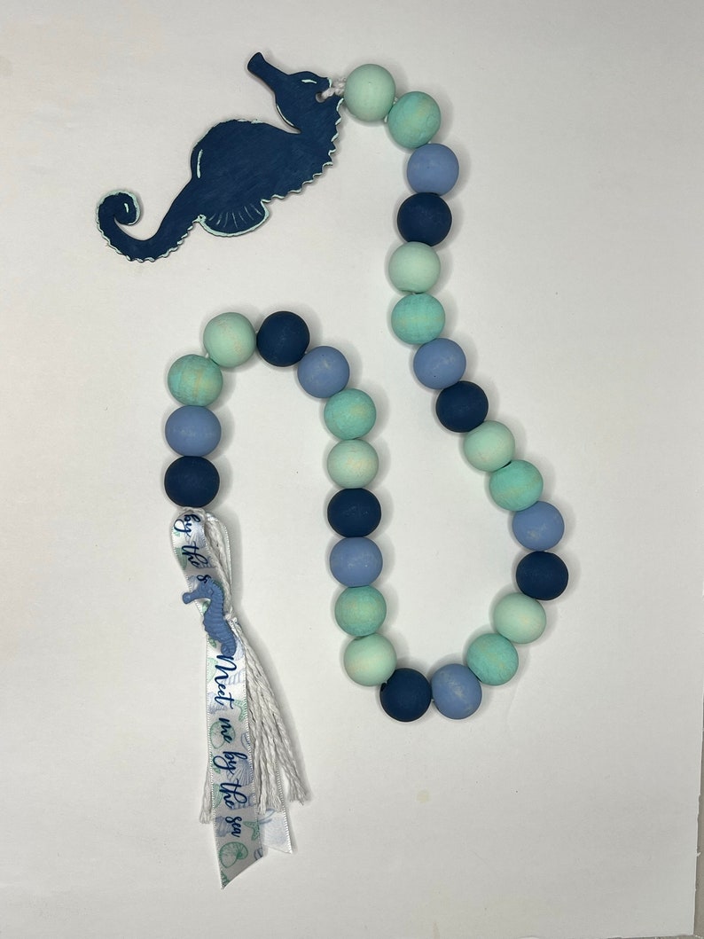 May include: A decorative wooden bead garland in shades of blue and green, featuring a navy blue seahorse cutout. A ribbon with the text "Meet me by the sea" is attached to the garland. The beads are approximately 1 inch in diameter.