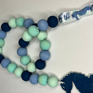 May include: A beaded garland with a nautical theme. The garland features wooden beads in shades of blue and green. A seahorse-shaped charm and a ribbon with the text "Meet me by the sea" complete the design.