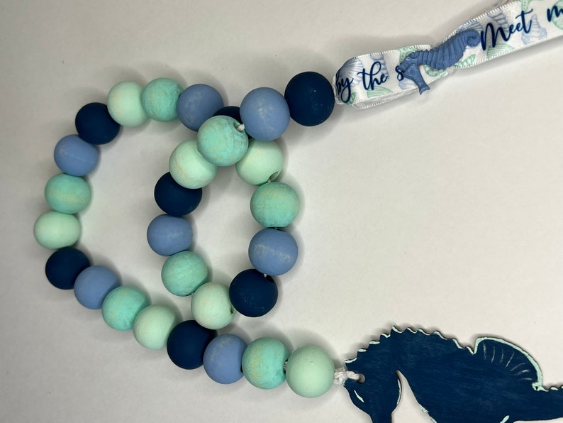 May include: A beaded garland with a nautical theme. The garland features wooden beads in shades of blue and green. A seahorse-shaped charm and a ribbon with the text "Meet me by the sea" complete the design.