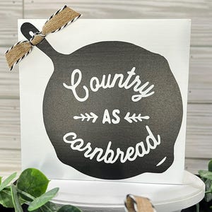 May include: A white wooden sign with a black skillet design and the phrase "Country as cornbread" in white script. A burlap ribbon with black and white trim is tied to the top left corner. The sign is on a white pedestal.