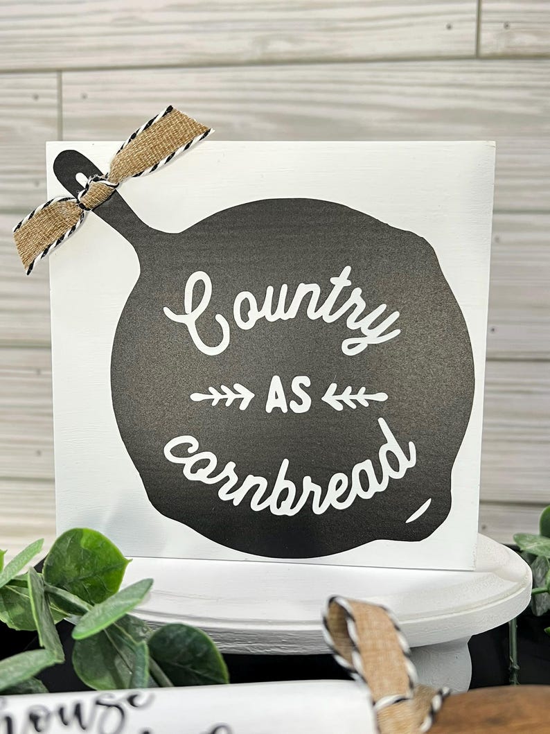 May include: A white wooden sign with a black skillet design and the phrase "Country as cornbread" in white script. A burlap ribbon with black and white trim is tied to the top left corner. The sign is on a white pedestal.