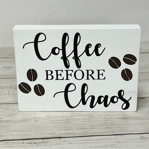 May include: White wooden sign with black lettering that reads "Coffee Before Chaos". The sign is decorated with brown coffee bean illustrations.