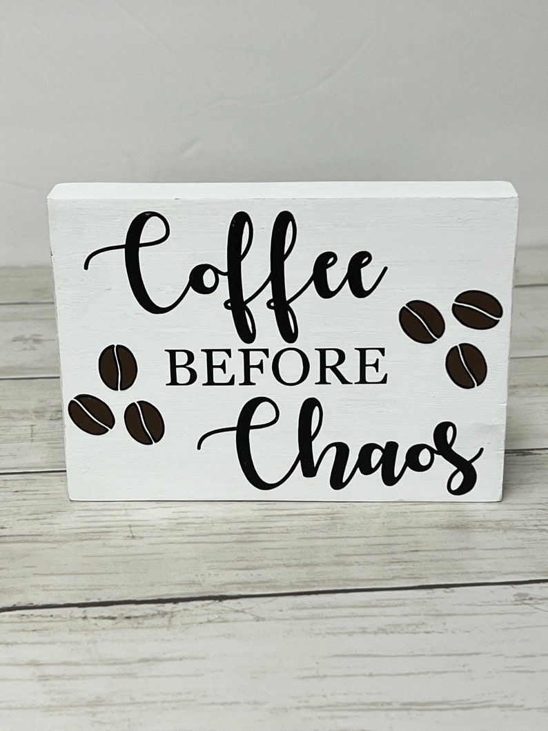 May include: White wooden sign with black lettering that reads "Coffee Before Chaos". The sign is decorated with brown coffee bean illustrations.
