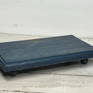 May include: A rectangular wooden display stand with a distressed blue paint finish. The stand has four rounded legs and a slightly raised edge around the top.