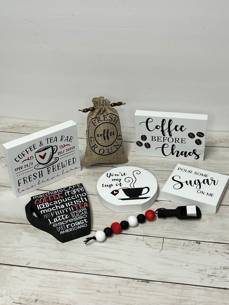 May include: A set of five white wooden signs with black lettering and coffee-themed sayings. The signs include "Coffee & Tea Bar", "Fresh Brewed", "You're my cup of", "Coffee Before Chaos", and "Pour Some Sugar On Me". There is also a small burlap bag with the words "Fresh Coffee Ground" printed on it, a black coffee mug with various coffee drink names printed on it, and a black and white beaded necklace.