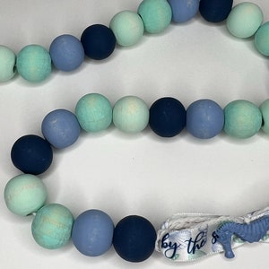 May include: A beaded garland featuring a mix of blue and green wooden beads. The garland includes a ribbon with the text "by the sea" and a seahorse charm. The beads are in various shades of blue and green.