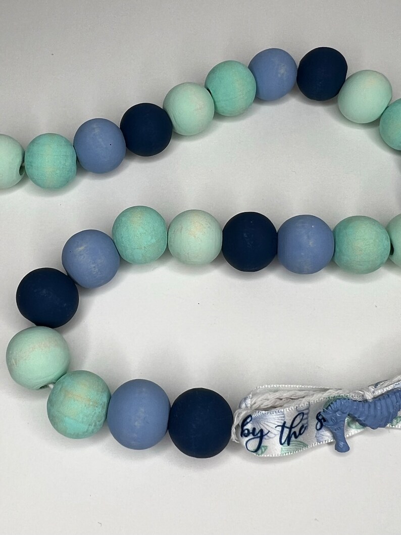 May include: A beaded garland featuring a mix of blue and green wooden beads. The garland includes a ribbon with the text "by the sea" and a seahorse charm. The beads are in various shades of blue and green.
