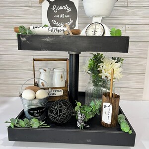 May include: A two-tiered black farmhouse-style tray with kitchen decor. The top tier holds a white scale, a sign that reads "Country as cornbread kitchen", and a rolling pin. The bottom tier has a bucket of eggs, a milk crate, and a jar of flowers.