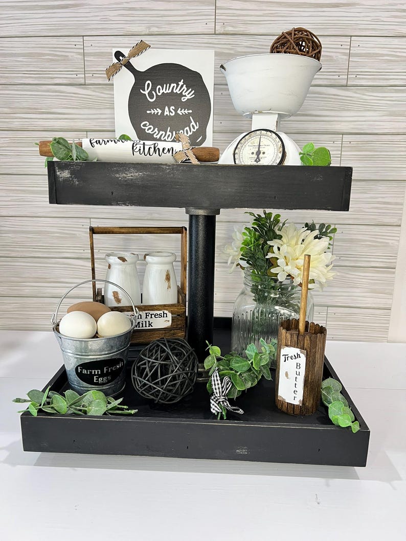 May include: A two-tiered black farmhouse-style tray with kitchen decor. The top tier holds a white scale, a sign that reads "Country as cornbread kitchen", and a rolling pin. The bottom tier has a bucket of eggs, a milk crate, and a jar of flowers.