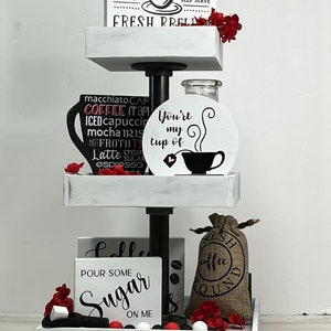 May include: A white tiered tray with three levels, decorated with coffee-themed signs and accessories. The top level has a sign that reads "Coffee & Tea Bar, Open 24/7, Self Serve, Fresh Brewed". The middle level has a sign that reads "You're my cup of" with a coffee cup and steam design. The bottom level has a sign that reads "Pour some Sugar on me" and a burlap bag with the words "Fresh Coffee Ground".