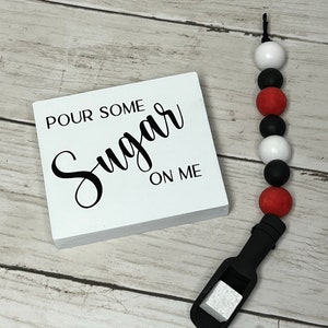 May include: White wooden sign with black text that reads "Pour Some Sugar On Me". A black silicone teething toy with red, white, and black beads is next to the sign.