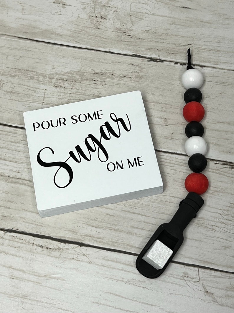 May include: White wooden sign with black text that reads "Pour Some Sugar On Me". A black silicone teething toy with red, white, and black beads is next to the sign.