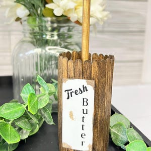 May include: A wooden butter churn with a label that reads "Fresh Butter." The churn is made of small wooden slats and has a wooden handle. The background includes a glass jar with white flowers and greenery.
