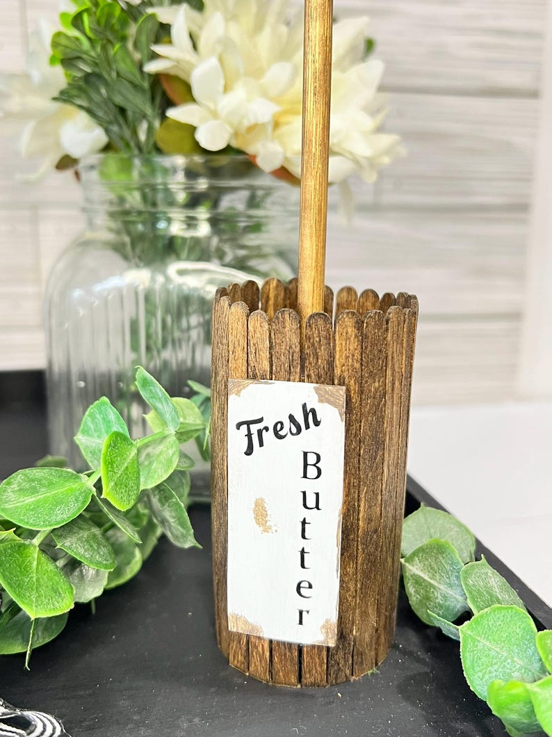 May include: A wooden butter churn with a label that reads "Fresh Butter." The churn is made of small wooden slats and has a wooden handle. The background includes a glass jar with white flowers and greenery.