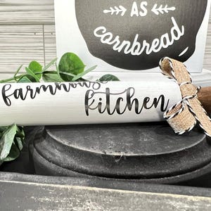 May include: A white rolling pin with the words "farmhouse kitchen" in black script. The rolling pin has wooden handles and is decorated with a burlap ribbon. A sign in the background reads "as cornbread".