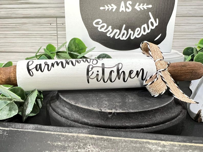 May include: A white rolling pin with the words "farmhouse kitchen" in black script. The rolling pin has wooden handles and is decorated with a burlap ribbon. A sign in the background reads "as cornbread".