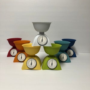 May include: A set of six colorful vintage-style kitchen scales with white faces and black numbers. The scales are arranged in a pyramid shape with a white bowl on top.