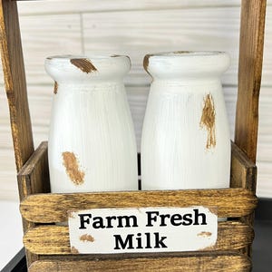 May include: A rustic wooden milk bottle holder with two white painted bottles. The bottles have brown distressed accents. The holder has a handle and a sign that reads "Farm Fresh Milk".