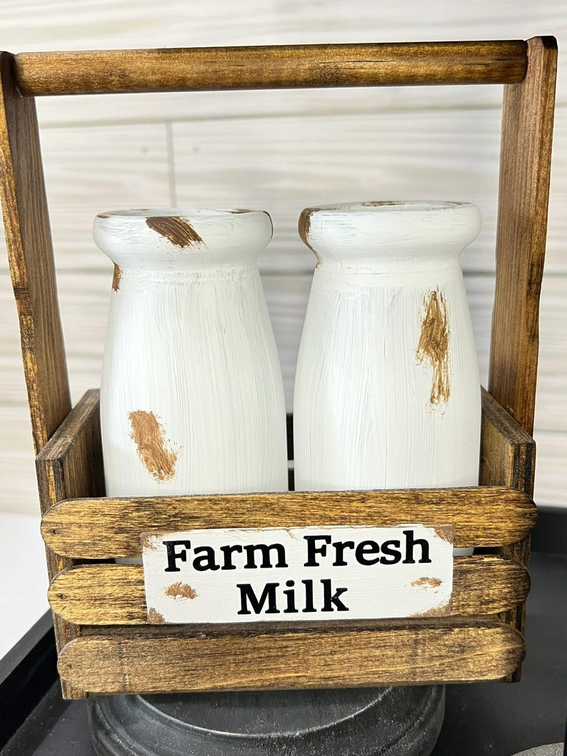 May include: A rustic wooden milk bottle holder with two white painted bottles. The bottles have brown distressed accents. The holder has a handle and a sign that reads "Farm Fresh Milk".