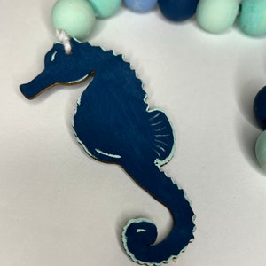 May include: A navy blue wooden seahorse charm with a light blue trim is attached to a strand of painted wooden beads in shades of blue and green. The beads are approximately 1 inch in diameter. The seahorse is a decorative element.