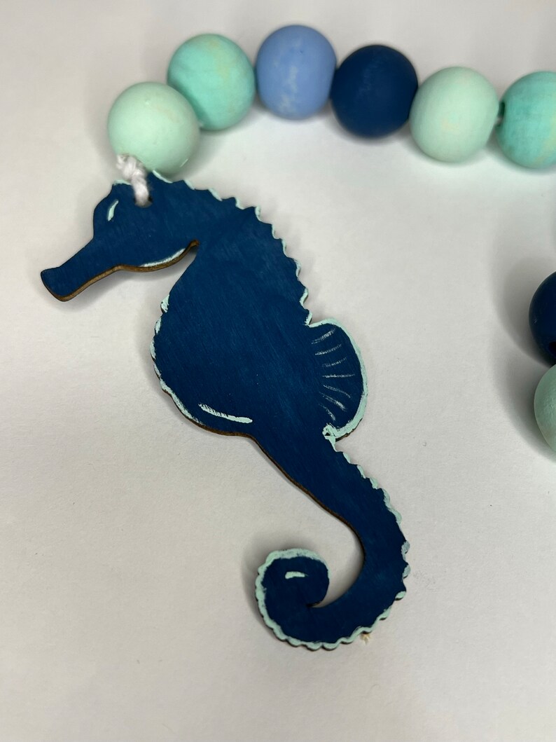 May include: A navy blue wooden seahorse charm with a light blue trim is attached to a strand of painted wooden beads in shades of blue and green. The beads are approximately 1 inch in diameter. The seahorse is a decorative element.