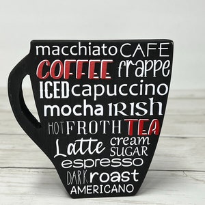 May include: Black wooden coffee cup shaped sign with white text listing different types of coffee and tea, including macchiato, frappe, iced cappuccino, mocha, latte, espresso, and americano.