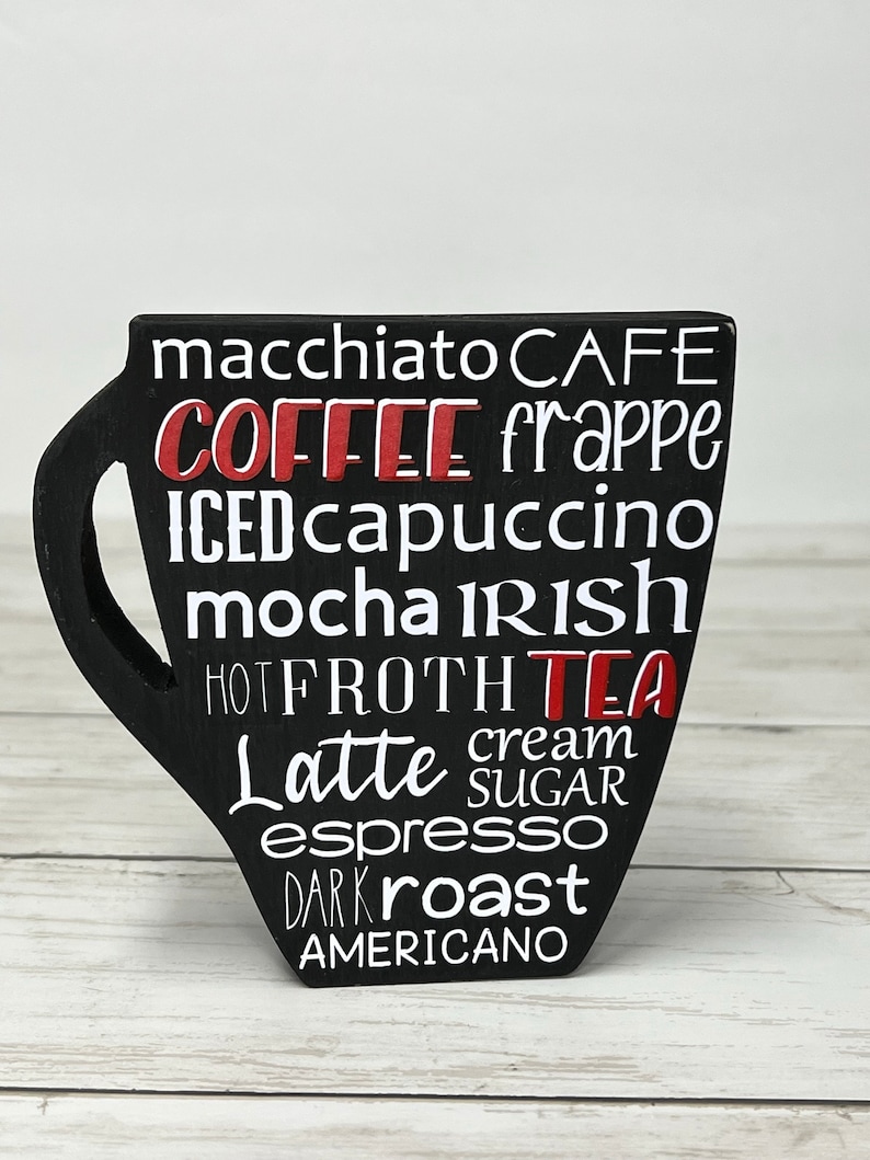 May include: Black wooden coffee cup shaped sign with white text listing different types of coffee and tea, including macchiato, frappe, iced cappuccino, mocha, latte, espresso, and americano.