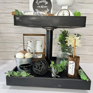 May include: A two-tiered black farmhouse-style tray with decorative items. The top tier holds a white scale, a sign that reads "Country as a Kitchen", and a wooden rolling pin. The bottom tier has a bucket of eggs, a wooden butter holder, and greenery.