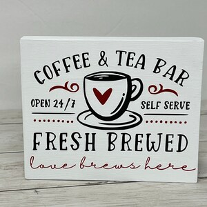 May include: White wooden sign with black text that reads "COFFEE & TEA BAR" with a stylized coffee cup and saucer with a red heart in the cup. The text "OPEN 24/7" and "SELF SERVE" are below the cup. The text "FRESH BREWED" is in larger black letters below the cup and the text "love brews here" is in smaller red letters below that.