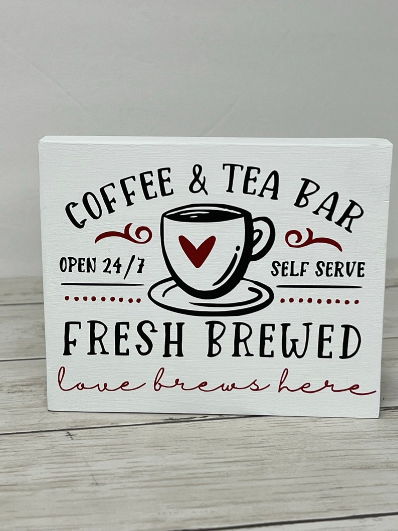 May include: White wooden sign with black text that reads "COFFEE & TEA BAR" with a stylized coffee cup and saucer with a red heart in the cup. The text "OPEN 24/7" and "SELF SERVE" are below the cup. The text "FRESH BREWED" is in larger black letters below the cup and the text "love brews here" is in smaller red letters below that.