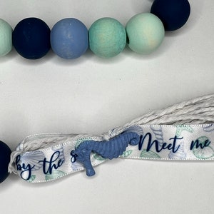 May include: A close-up of a baby teething necklace with a variety of blue and green beads. The necklace has a ribbon with the text "Meet me by the sea" and a blue seahorse charm.