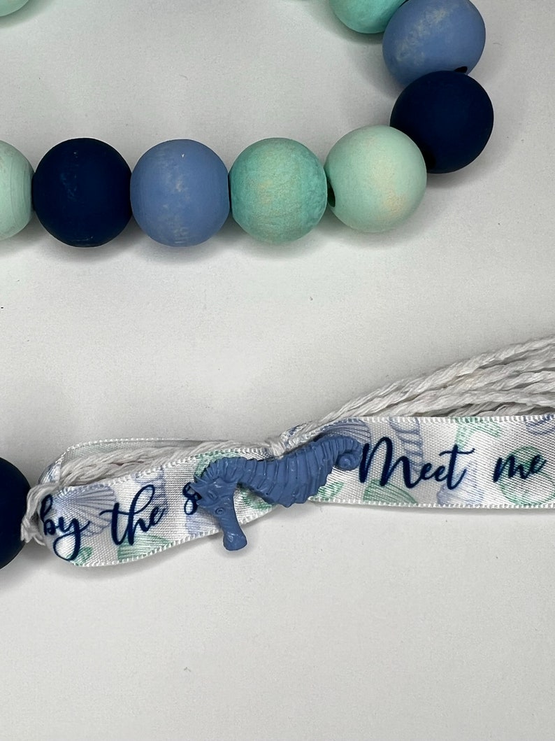 May include: A close-up of a baby teething necklace with a variety of blue and green beads. The necklace has a ribbon with the text "Meet me by the sea" and a blue seahorse charm.
