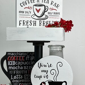 May include: A white tiered tray with a sign that reads "Coffee & Tea Bar Open 24/7 Self Serve Fresh Brews". The tray is decorated with a black coffee cup with the words "macchiato, CAF, COFFEE, frapi, ICED, cappuccin, mocha, IRIS, HOT, FROTH, T, Latte, crea, sugar, espresso, DARK, roast, CANO" written on it. There is also a white round sign with the text "You're my cup of" and a black coffee cup with a heart on the tea bag.