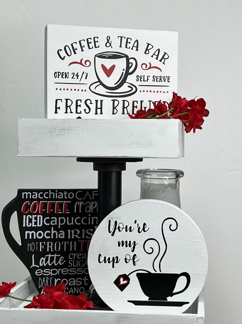 May include: A white tiered tray with a sign that reads "Coffee & Tea Bar Open 24/7 Self Serve Fresh Brews". The tray is decorated with a black coffee cup with the words "macchiato, CAF, COFFEE, frapi, ICED, cappuccin, mocha, IRIS, HOT, FROTH, T, Latte, crea, sugar, espresso, DARK, roast, CANO" written on it. There is also a white round sign with the text "You're my cup of" and a black coffee cup with a heart on the tea bag.