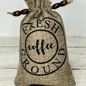 May include: A brown burlap bag with a black drawstring and wooden beads. The bag has a black circle with the text "FRESH GROUND COFFEE" printed on it.