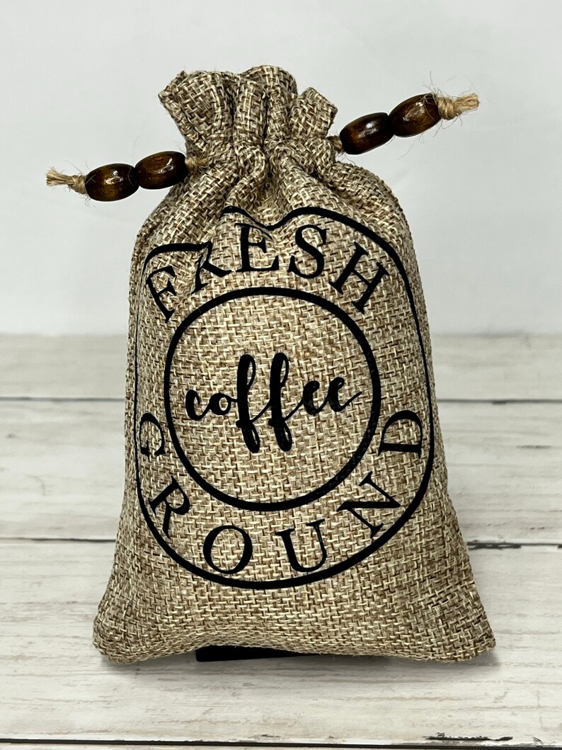 May include: A brown burlap bag with a black drawstring and wooden beads. The bag has a black circle with the text "FRESH GROUND COFFEE" printed on it.