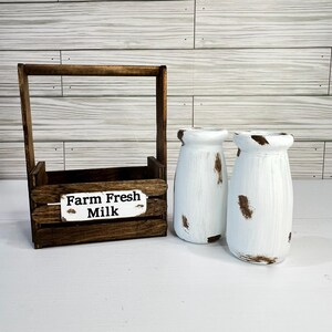 May include: A rustic wooden milk crate with a handle and a sign that reads "Farm Fresh Milk." Two white, distressed milk bottles with brown accents sit beside the crate. The items are arranged on a white surface.