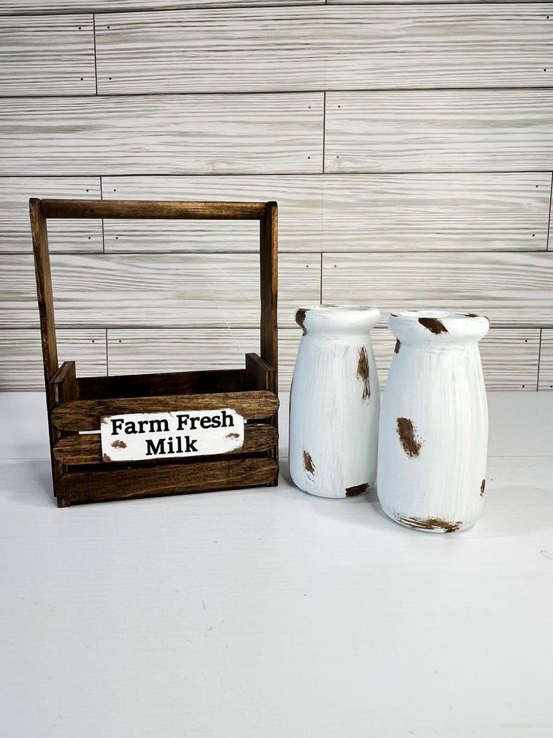 May include: A rustic wooden milk crate with a handle and a sign that reads "Farm Fresh Milk." Two white, distressed milk bottles with brown accents sit beside the crate. The items are arranged on a white surface.