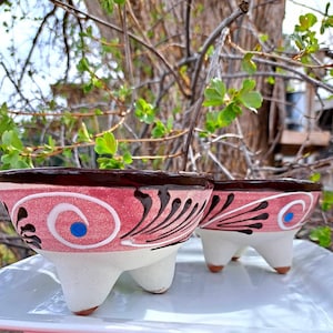 May include: Two terracotta bowls with three legs each. The bowls are painted in pink, white, and brown with a blue swirl design.