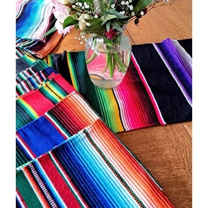 May include: A set of colorful striped table runners, with a black and purple runner in the foreground. The runners are made of a woven fabric and have a traditional Mexican design.