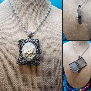 May include: A silver chain necklace with a silver locket pendant. The locket is shaped like a book and has a white floral design on the front.